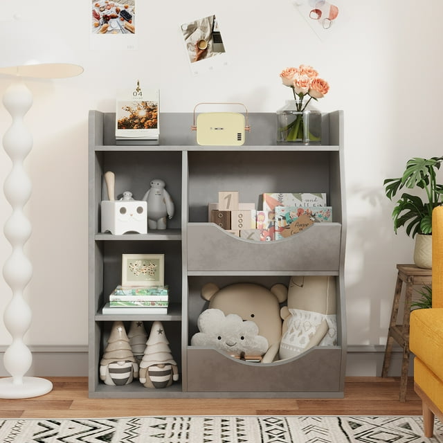 gabinete de almacenamiento de juguetes, estantería y estantería para niños, estante múltiple con organizador de cubos para almacenamiento de libros y juguetes vista frontal de mueble gris con tres compartimentos principales y dos cubos inclinables frontales; estantes laterales con libros, peluches y cajas; tablero de partículas con bordes redondeados y accesorio antivuelco, colocado en habitación infantil junto a lámpara de pie y planta. A menudo usado también como estantería baja para guardar ropa de muñecas o material de manualidades.