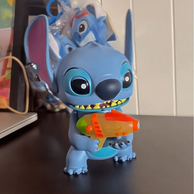 figuras de acción de lilo & stitch para niños, muñecos de stitch, juguete musical con sonido ligero, regalos — figura de Stitch de plástico de about 16cm, vista frontal centrada; cuerpo azul con orejas grandes y ojos negros, sostiene un accesorio tipo arma naranja y verde, dientes pintados visibles y acabado mate; fondo interior difuminado con otros muñecos. A menudo usada como figura de exhibición en estantes de colección.