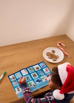 Child in a Santa hat colors a blue holiday activity sheet at a wooden table with gingerbread cookies, a candy cane and a green marker.