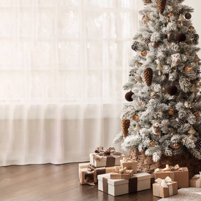 Snow frosted Christmas tree with pinecone and owl ornaments and wrapped gifts on a wooden floor in a bright living room.