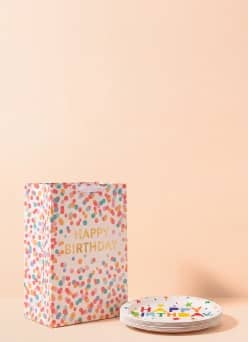 Happy Birthday gift bag with colorful confetti pattern next to stacked white plates with multicolor party designs.