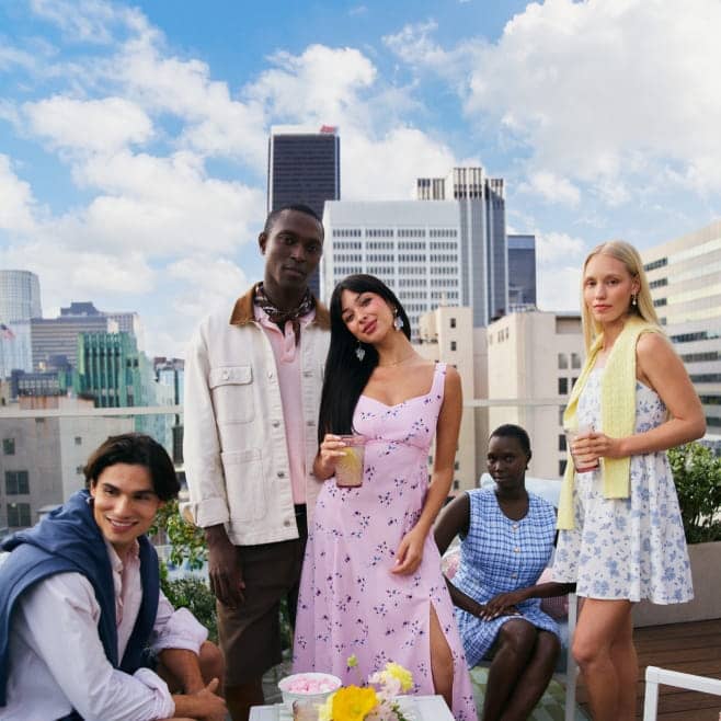 Five friends pose on a rooftop patio in front of a city skyline, wearing pastel spring outfits and holding drinks around a small table with flowers.