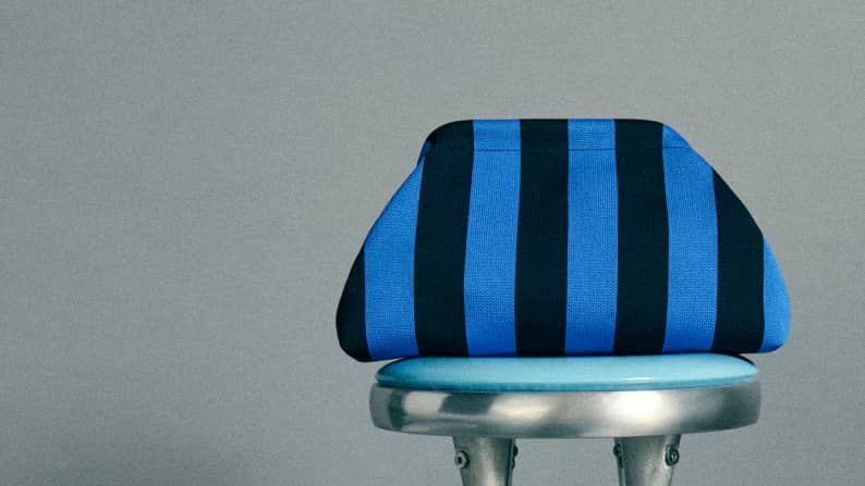 A blue and black striped oversized clutch bag sits on top of a metal stool against a gray background.