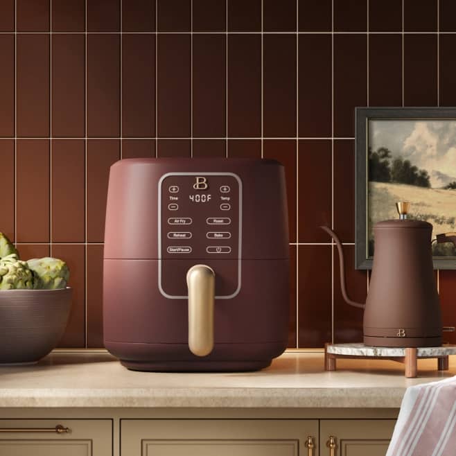 Maroon air fryer with digital controls on a kitchen counter beside a bowl of artichokes and a matching carafe.