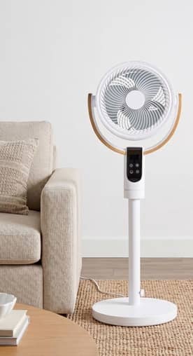 A white fan on a wood floor, next to a beige sofa.