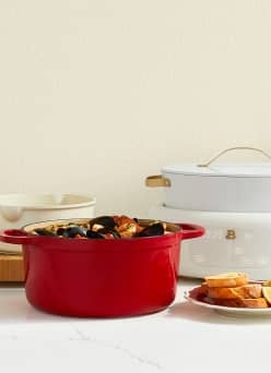 A red cast iron pan is on a white countertop, next to a white slow cooker and a plate of bread.