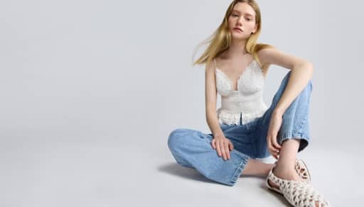 Woman with long blonde hair wearing a white lace trim camisole top and light blue jeans seated on the floor against a light gray background.