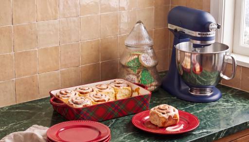 Tray of cinnamon rolls in a red plaid dish on a green counter with a blue stand mixer, red plates and a jar of holiday cookies.