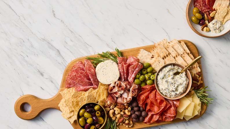 Wooden board with sliced meats, cheeses, crackers, olives, nuts and herb dip, with a smaller plate of similar snacks nearby.