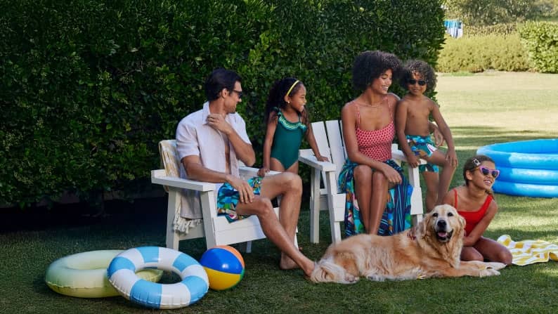 A family of five wearing colorful swimsuits and swim shorts is sitting outside together surrounded by pool toys and a golden retriever.