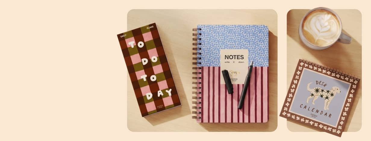 A planner, notebook, calendar, pen, and coffee mug on wood table.