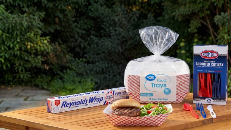 Reynolds Wrap foil, a bag of paper food trays, and a box of cutlery on a wooden table, with a burger and salad in a paper tray.