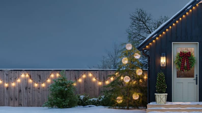 Snowy outdoor shot of A-frame house and fence decorated with string lights, a Christmas wreath & oversized ornaments in the trees.