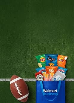Against the background of a football field, snacks, drinks and condiments overflow from a Walmart Business shopping bag, which sits beside a football.