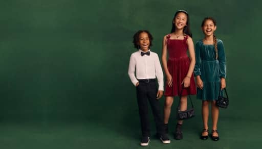 A boy wearing formal clothes and a bowtie, a girl wearing a red dress, and a girl wearing a blue green dress stand in front of a green background.
