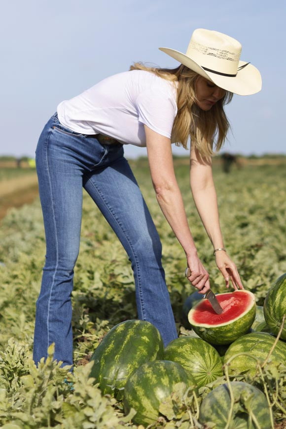 A woman wearing a sun hat in a field is leaning over to cut into a ripe watermelon on top of a small pile of whole watermelons.