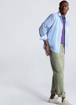 A man poses in a light blue button-up over a striped shirt with khaki pants and white sneakers.