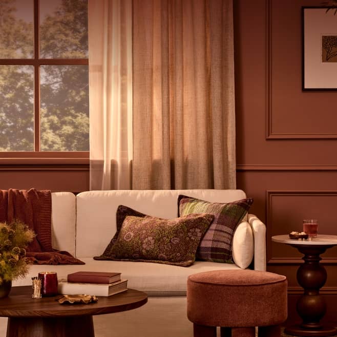 Cozy living room with light sofa, patterned pillows, round wood coffee table with books and candle, and ottoman in warm autumn colors.