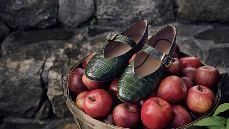 Pair of green Mary Jane flats with buckle straps, styled over a basket filled with red apples.