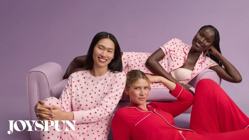 Three women smiling and lounging against a purple background, wearing cherry print pajamas and a bright red pajama set.