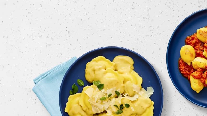 A blue plate with ravioli, cream sauce, herbs, and shaved Parmesan next to a blue plate with pasta and red sauce on a white countertop.