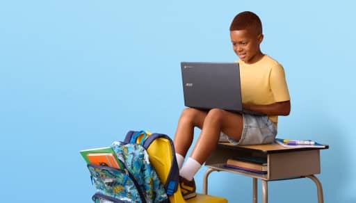 Child sitting on school desk using laptop, with backpack, notebooks and school supplies nearby.