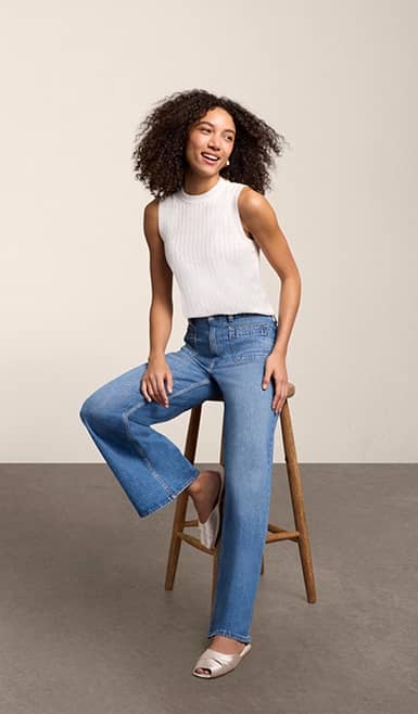 A woman wearing wide leg jeans and a white tank top is pictured sitting on stool against a beige backdrop.