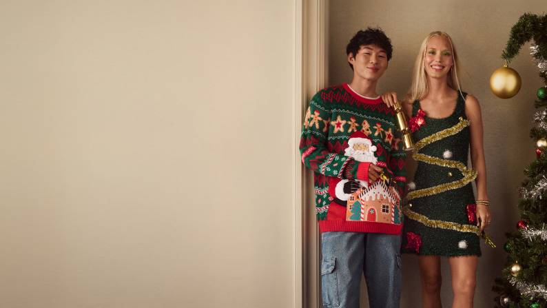 A young man wearing a Santa sweater and young woman wearing Christmas tree dress are pictured in a home next to a whimsical Christmas tree.