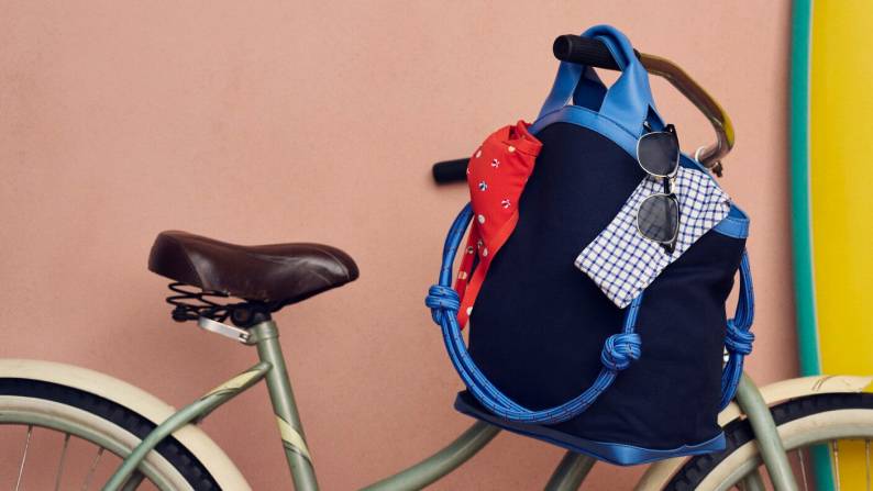 Navy tote with rope handles hanging on a bicycle, styled with sunglasses and other accessories against a pastel backdrop.