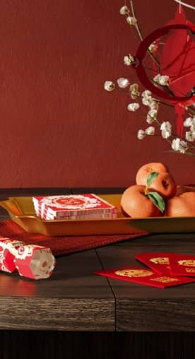 A festive tabletop scene with a red hanging lantern, blossoming branches, tangerines, a red party cracker, napkins and red envelopes.