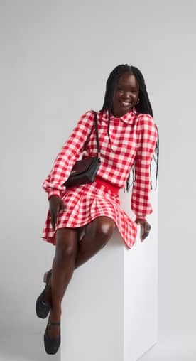 A woman is shown wearing a red plaid jacquard collared sweater with long puff sleeves with a matching pleated mini sweater skirt from Free Assembly.