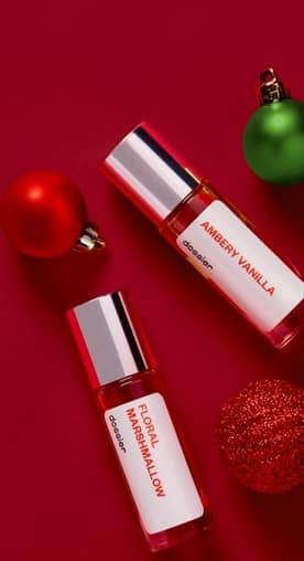 Two mini Dossier perfume bottles labeled “Floral Marshmallow” and “Ambery Vanilla” are arranged around red and green ornaments.