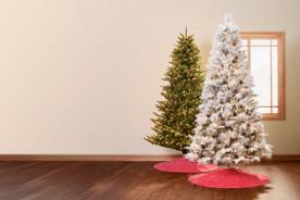 Image of two Christmas trees in a room. One is a flocked tree with lights and a red skirt, and the other is a green pre-lit tree with a red skirt.
