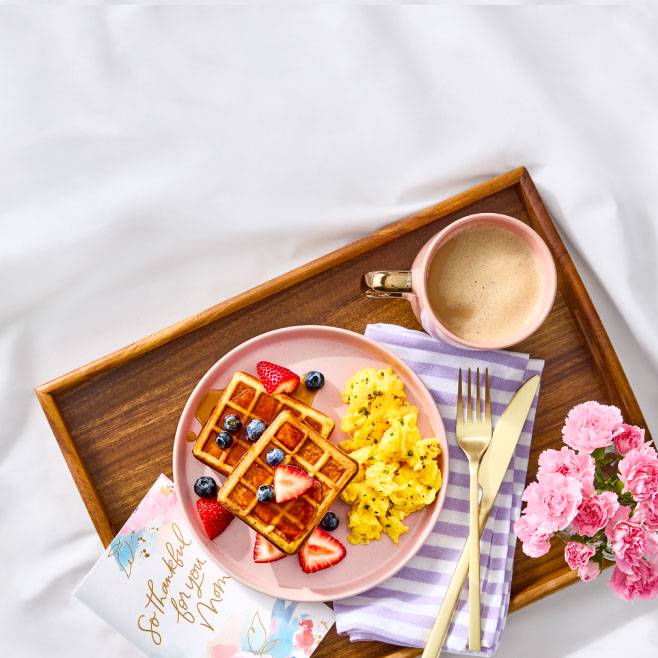 A wooden tray with a greeting card, flowers, coffee, and a plate of scrambled eggs and waffles topped with syrup and fresh berrie