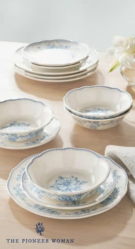 A set of white ceramic dinnerware with a charming light blue floral design is set on a light wooden table with a napkin and a vase of flowers.