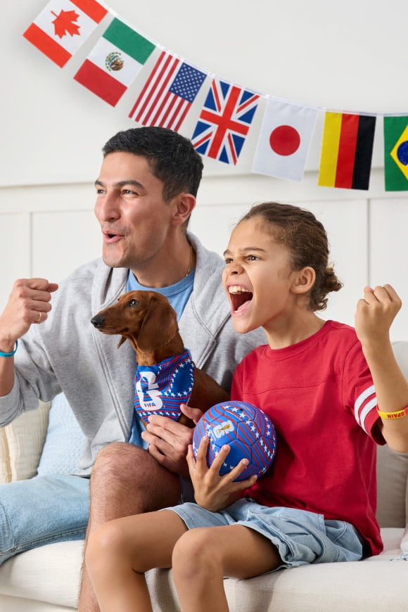 A multi-country flag banner, a TV with a World Cup display, a twelve pack of Coke, two cans of Celsisus, paper plates and napkins and plate of tacos.