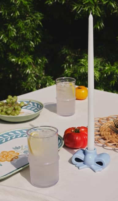 Outdoor summer tablescape with pastel dishware, a tall white candle, a tomato & citrus drinks styled on a white table in sunlight.