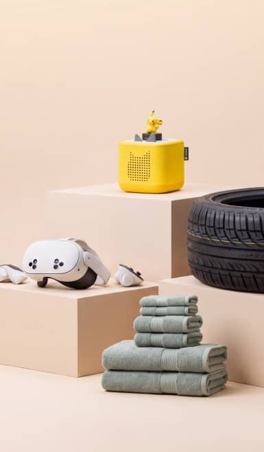 Yellow cube speaker, white virtual reality headset, folded green towels and a black car tire on minimalist podiums.