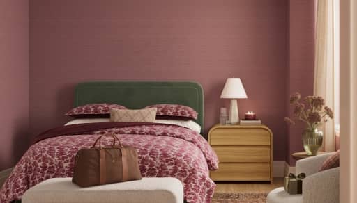 Cozy bedroom with green headboard, patterned burgundy bedding, nightstand with lamp and candle, and leather travel bag on bench.