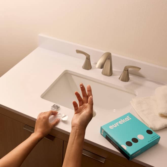 Eurekar contact lenses box on a bathroom counter as a person prepares a lens from a blister pack near the sink.
