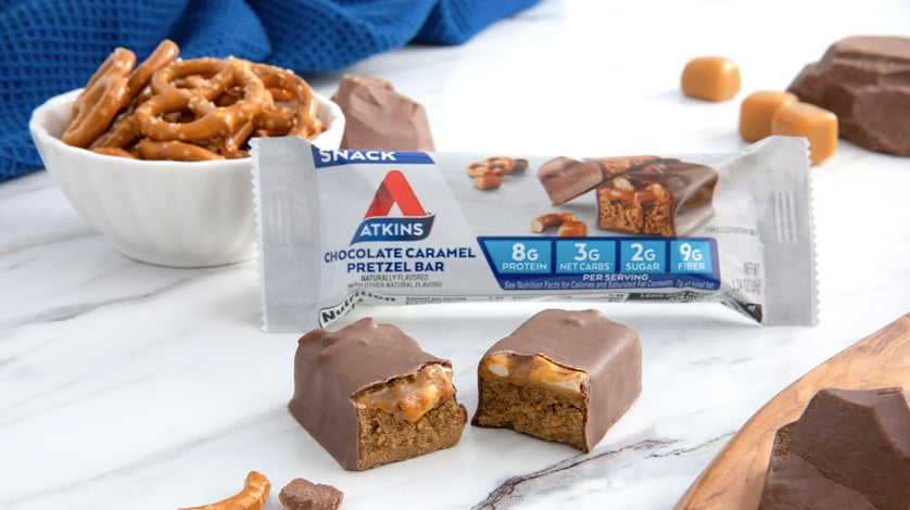An Atkins snack bar in its package next to chocolate covered pretzels in a bowl