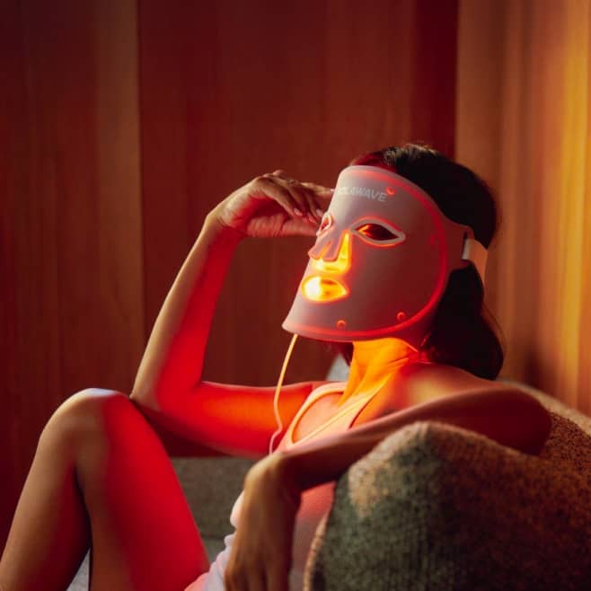 Person reclining on a chair wearing an illuminated LED skincare mask in a warm, low-lit room.