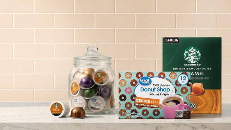 A glass container full of mixed coffee pods next to Great Value donut shop coffee pods and Starbucks caramel coffee pods on a marble countertop.