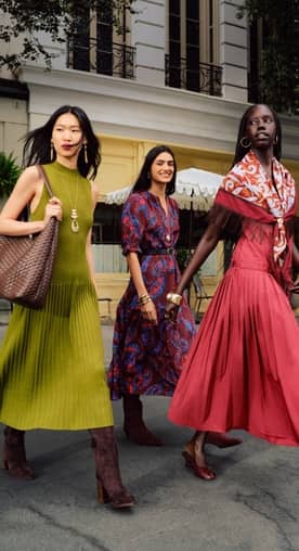 Three adults walk outdoors wearing colorful long dresses and boots, with one carrying a woven bag and another wearing a patterned scarf.