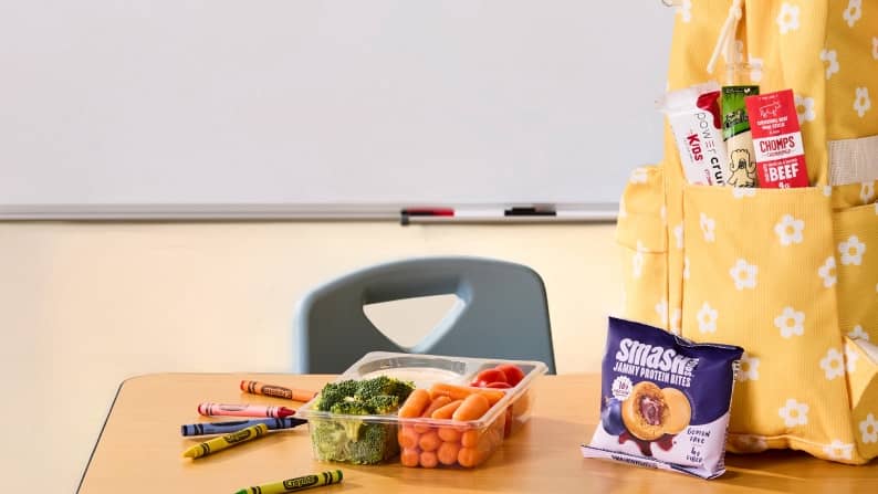A small tray of carrots, tomatoes, broccoli, and dip on a desk next to Smash jammy protein bites and a backpack with a Chomps stick and other snacks.
