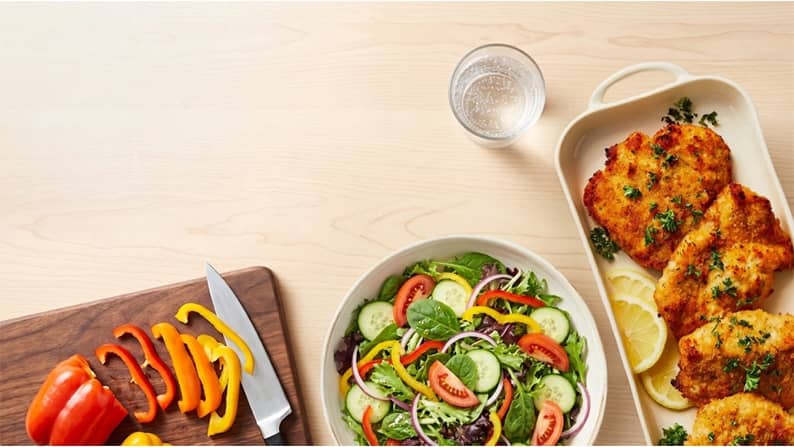 A cutting board with bell peppers, a salad, a beverage in a glass, and a tray of chicken tenders.