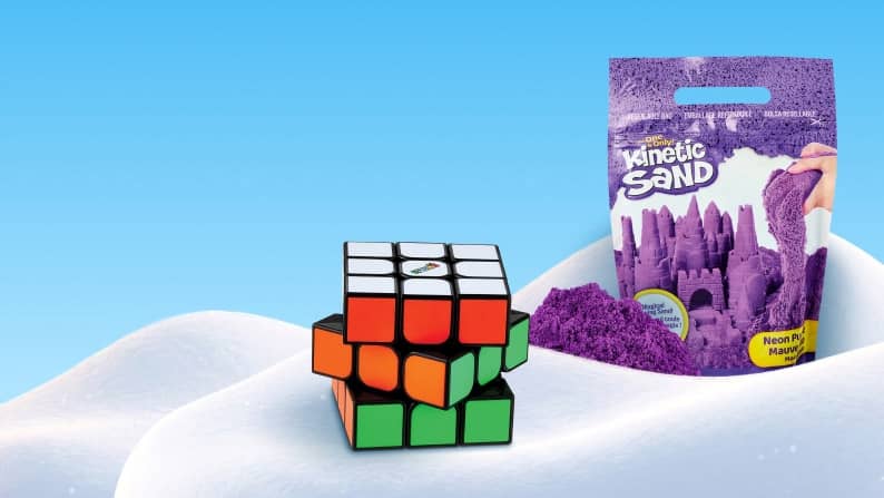 Kinetic Sand purple sand bag with sculpted sandcastles beside a multicolored puzzle cube on a smooth white surface.