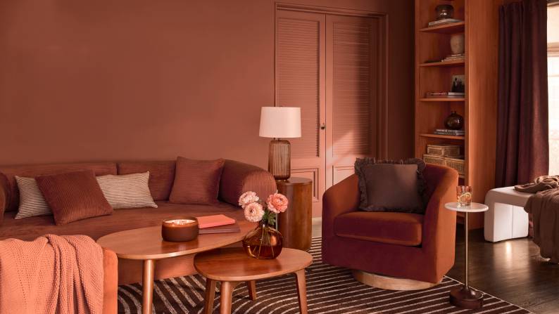 A terracotta living room with a ribbed sofa, velvet swivel chair and wooden nesting tables. A striped rug and built-in bookshelf are in the back.