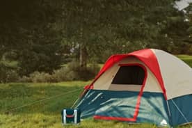 A blue tent, camp chair, and cooler on a green lawn next to a fire pit.