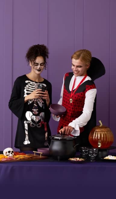 Two adults in skeleton and vampire costumes serve food from a cauldron on a Halloween snack table with skulls and a pumpkin drink dispenser.
